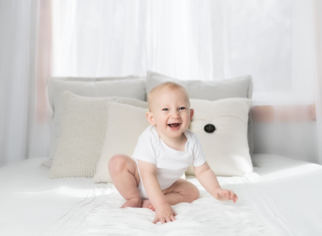Offerings Cute baby sitting and smiling on a white bed in a bright, airy bedroom.