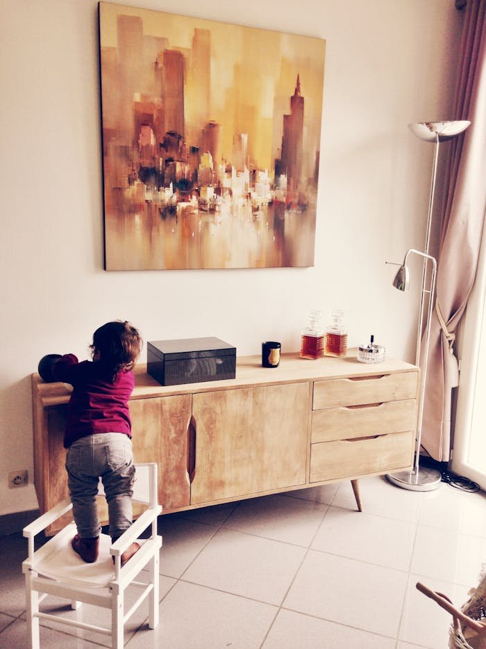 Child standing on chair reaching a wooden cabinet in a modern living room with abstract painting.